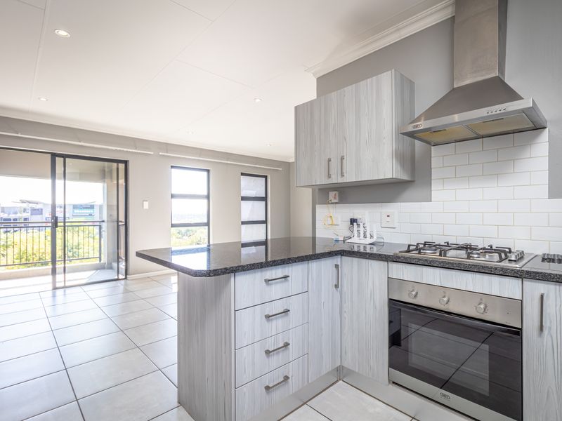 Modern open-plan kitchen featuring light wood-grain cabinetry, granite countertops, and integrated appliances, flowing into a bright living area with access to a private balcony.
