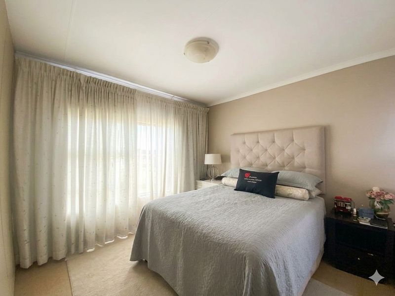 A well-maintained bedroom featuring neutral wall tones, carpeted flooring, and large windows dressed with floor-to-ceiling curtains.