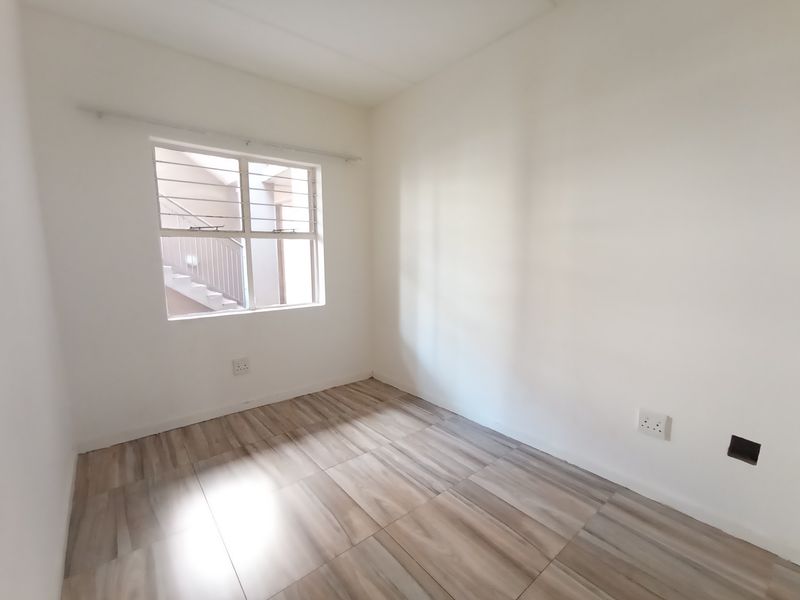 A bright, empty bedroom featuring modern wood-look laminate flooring, a window with security bars, and neutral white walls.