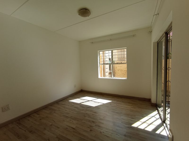 A bright, empty bedroom featuring modern laminate flooring, a sliding glass door with security gate, and a window with security bars.