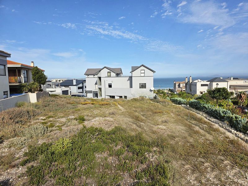 A vacant residential plot featuring natural coastal vegetation with surrounding established homes and ocean views in the distance.