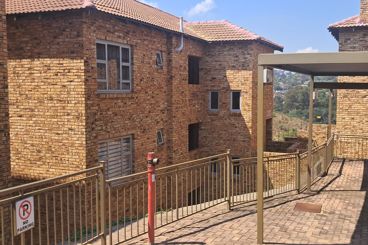 Exterior view of a face brick townhouse complex featuring a paved patio area, metal balustrades, and a covered carport structure.
