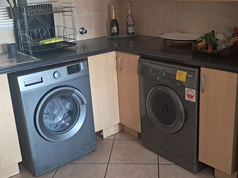 A functional laundry area featuring laminate cabinetry, dark countertops, a stainless steel sink, and tiled flooring with space for under-counter appliances.