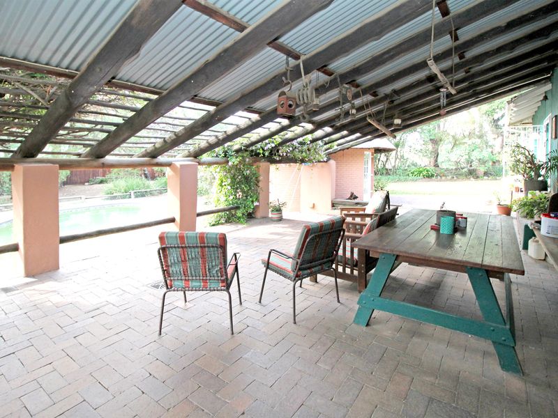 A covered outdoor patio area featuring a corrugated metal roof, timber support beams, and brick paving, overlooking the garden and pool area.