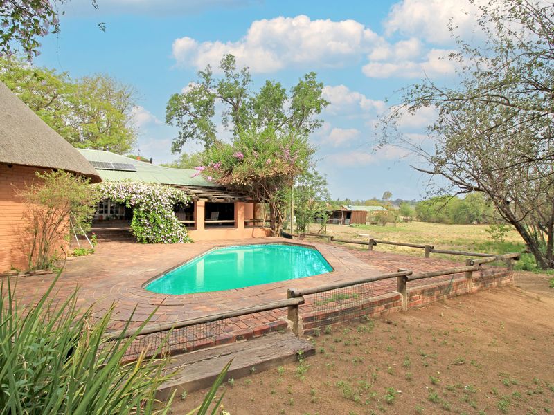 Outdoor pool area featuring a brick-paved deck, timber fencing, and a covered patio attached to a thatch-roofed farm dwelling.