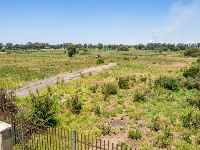 An aerial view of a large, undeveloped residential land parcel featuring overgrown vegetation, a paved access road, and perimeter fencing.