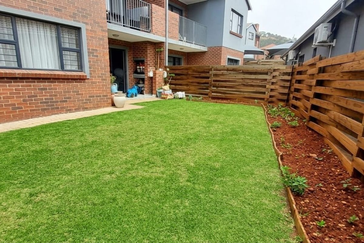 A well-maintained rear garden featuring a neat lawn, timber fencing, paved patio area, and a built-in braai against the brick apartment facade.