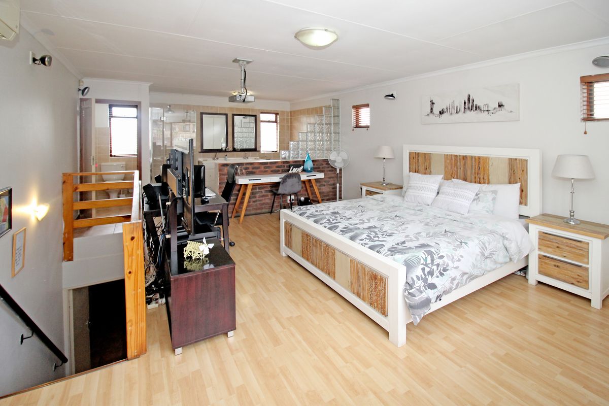 Spacious open-plan bedroom featuring laminate flooring, an integrated en-suite bathroom with glass block accents, and a staircase leading to the lower level.