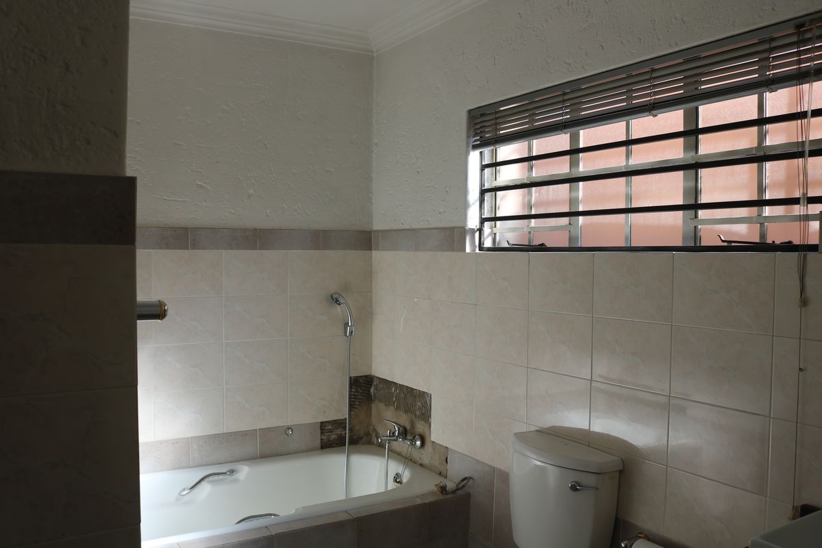 A bathroom featuring a built-in bathtub with shower mixer, toilet, and window with security bars, currently requiring tile repairs.