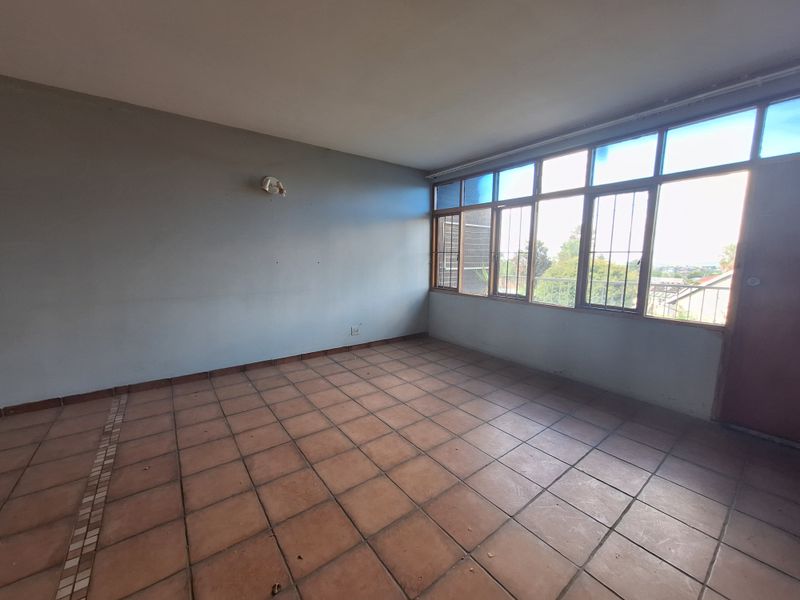 An empty living room featuring terracotta floor tiles and a large window bank with security bars, offering natural light and views.