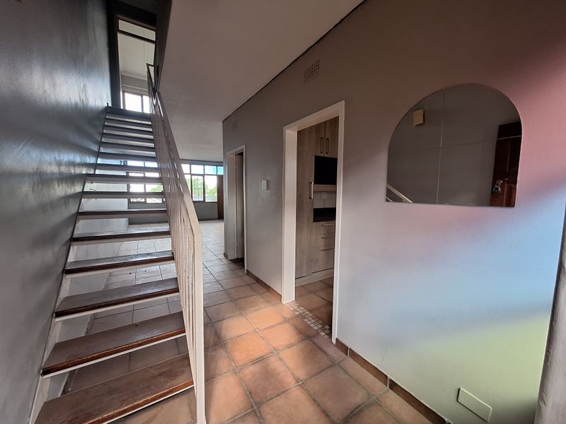 A spacious hallway featuring a staircase with metal railings, terracotta-style floor tiles, and access to adjacent rooms with built-in cabinetry.
