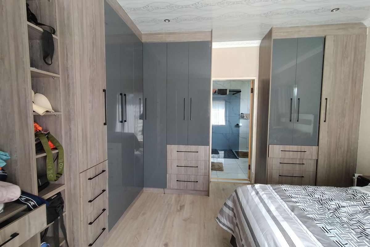 A well-appointed bedroom featuring extensive modern built-in cupboards with a two-tone finish, laminate flooring, and recessed lighting, with an adjoining bathroom visible.