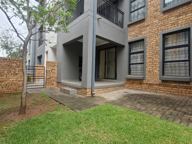 Rear view of the apartment featuring a private garden, paved patio area, and a covered balcony with modern face brick and grey plaster finishes.