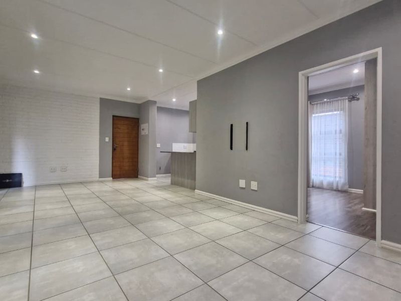 Spacious open-plan living area featuring neutral grey walls, a white brick feature wall, tiled flooring, and recessed downlighting, leading into the kitchen and adjacent rooms.