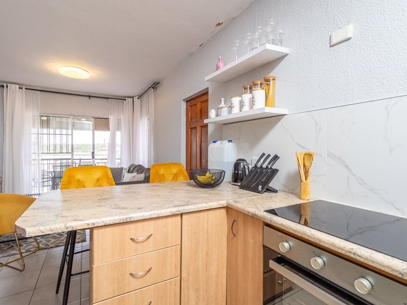Open-plan kitchen featuring light wood cabinetry, marble-effect splashback, and integrated appliances, flowing into a living area with access to a balcony.