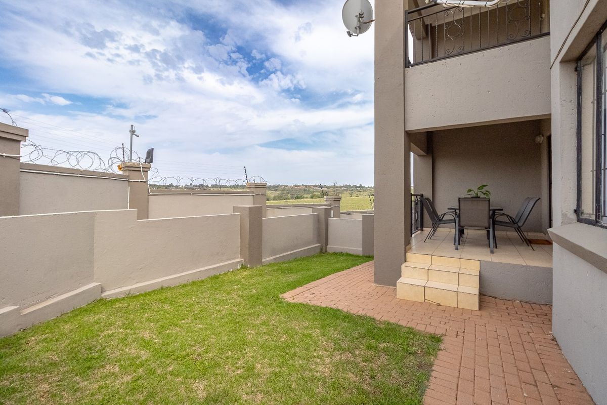 Rear exterior view featuring a paved patio area, small lawn, and perimeter wall with security fencing, adjacent to the building's lower level.