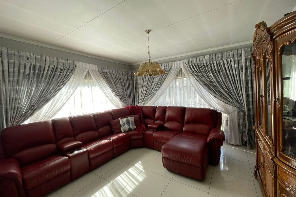 A spacious living room featuring large windows with layered curtains, tiled flooring, and a central pendant light fixture.