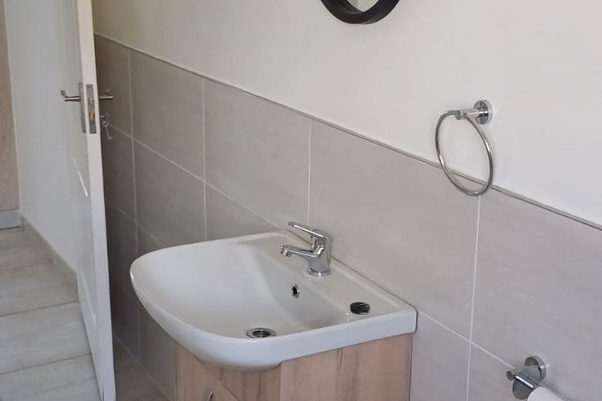 A clean and modern bathroom featuring a wood-finish vanity cabinet, ceramic basin, chrome fixtures, and neutral wall and floor tiling.