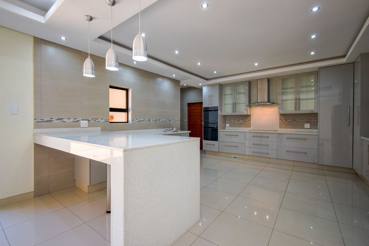 Modern kitchen featuring a large stone-topped breakfast island, sleek cabinetry, built-in oven, extractor fan, and polished floor tiles with recessed ceiling lighting.