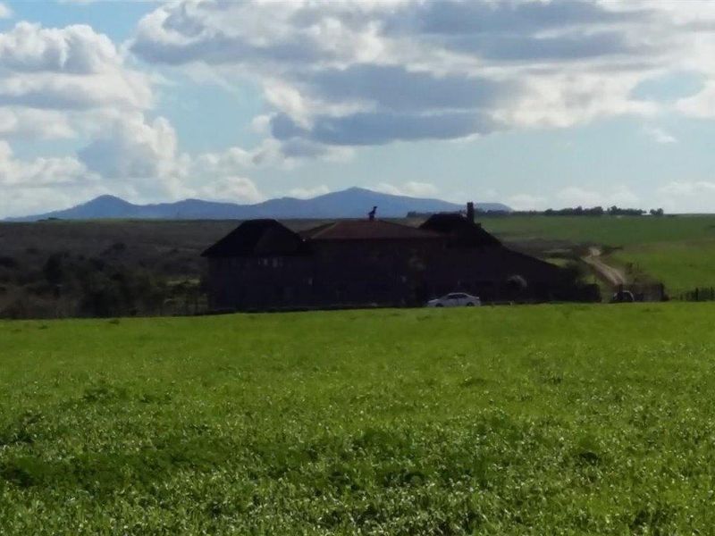 A distant view of a farmhouse set within expansive green fields, featuring a traditional pitched roof and chimney against a mountain backdrop.