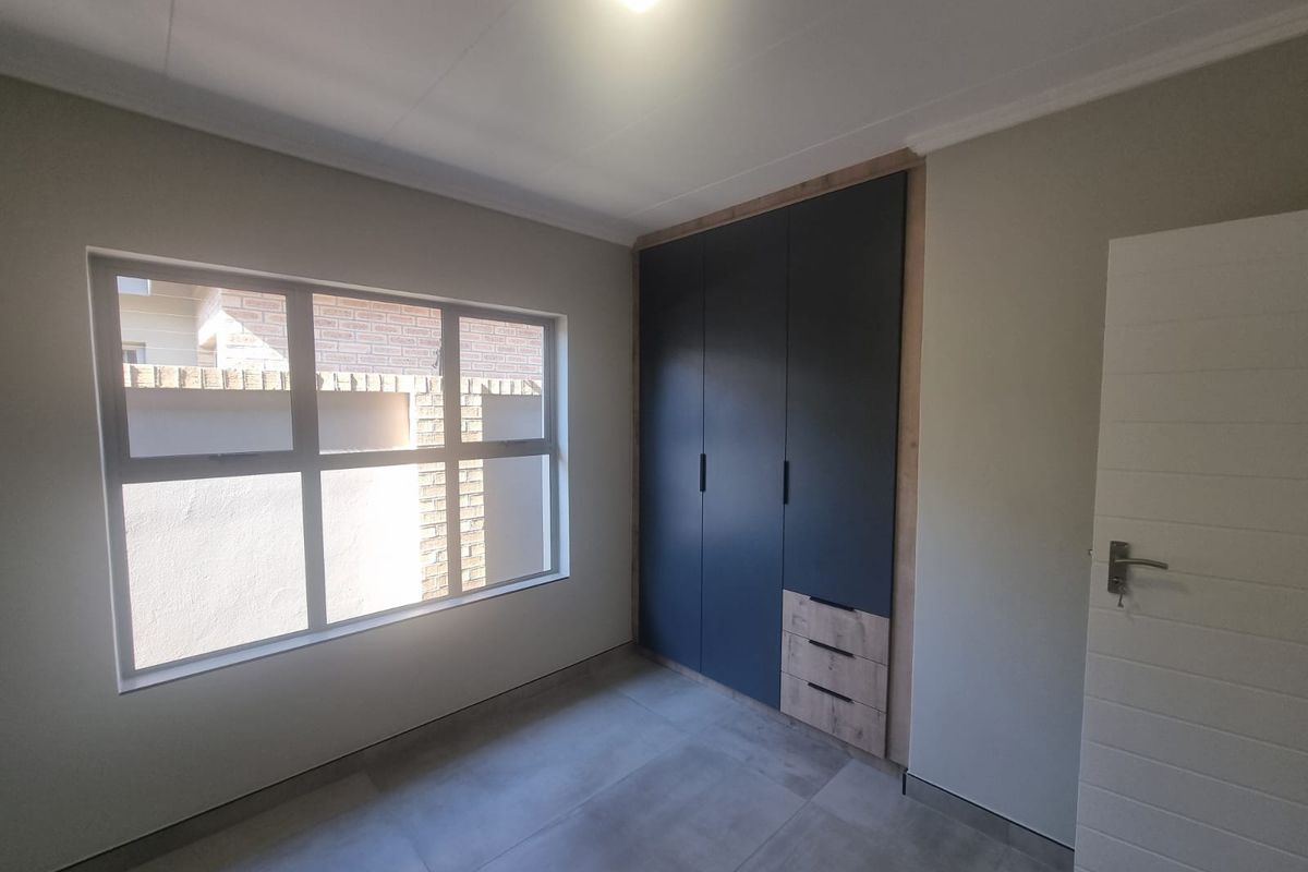 A bright, modern bedroom featuring neutral tiled flooring, a large window, and contemporary built-in cabinetry with a dark finish and wood-grain accents.