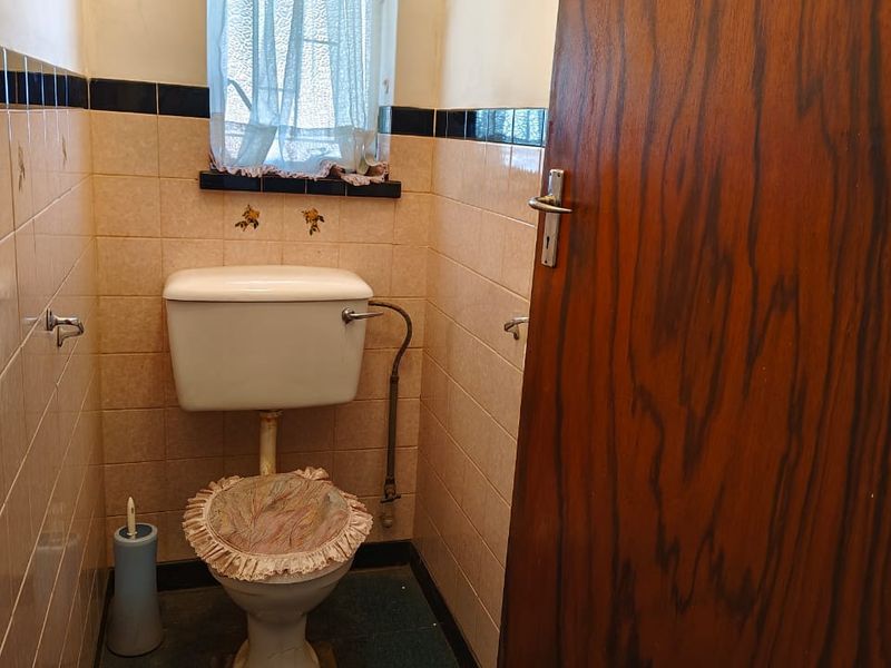 A compact, dated bathroom featuring original wall tiles with decorative accents, a standard toilet, and a wooden door.