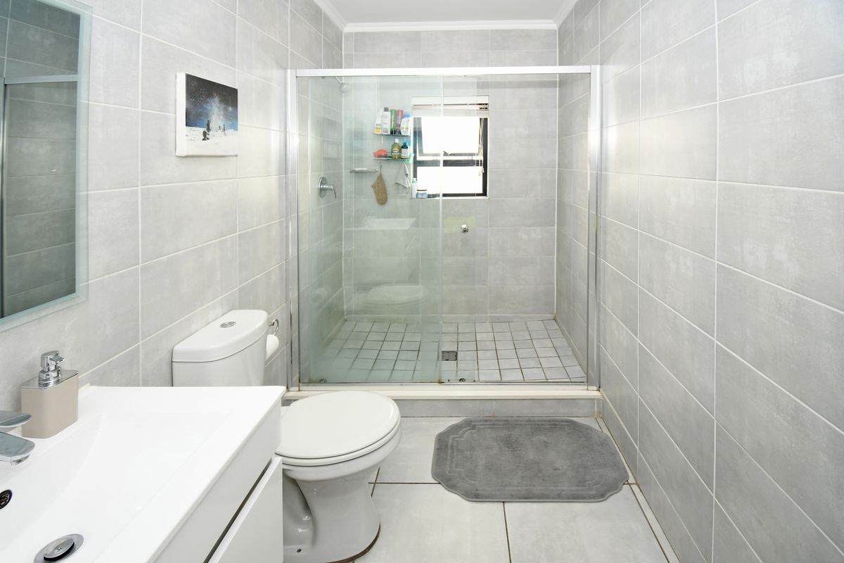 A modern bathroom featuring full-height grey wall tiling, a glass-enclosed shower, a white vanity basin, and a standard toilet.