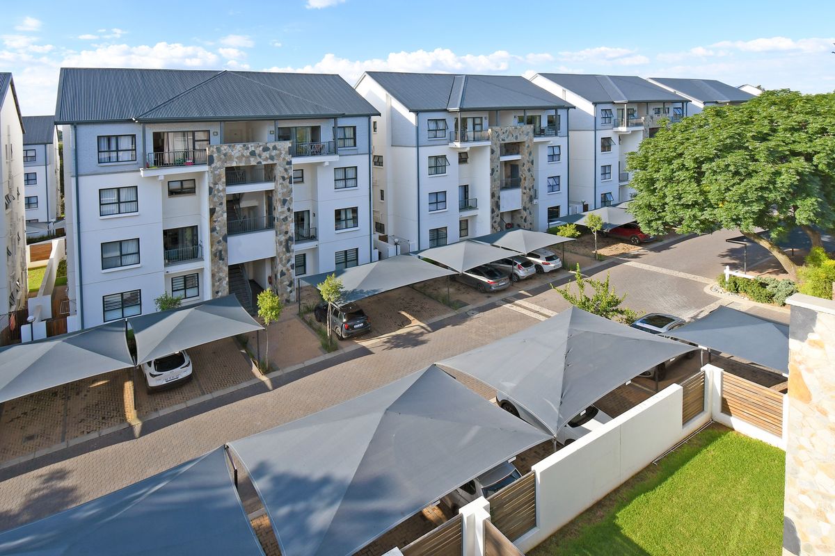 An elevated view of a modern apartment complex featuring multi-story residential buildings, paved parking areas with shade cloth carports, and landscaped grounds.