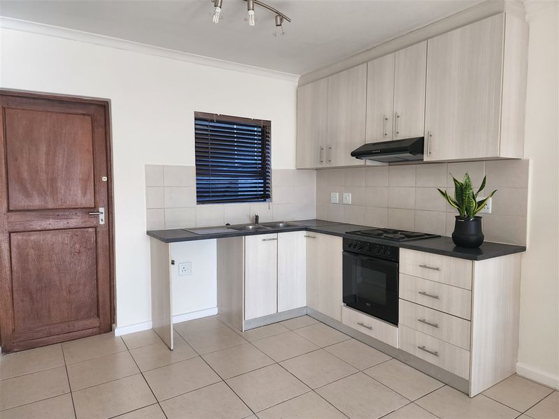 A functional kitchen featuring light wood-grain cabinetry, dark countertops, built-in oven and hob, tiled backsplash, and neutral floor tiles.