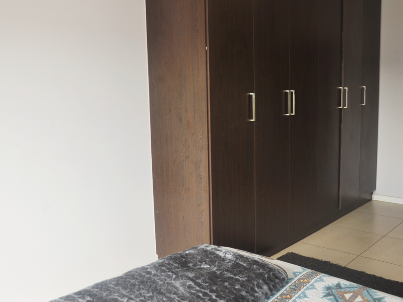 A bedroom featuring dark wood built-in cupboards and neutral tiled flooring, leading into a hallway.