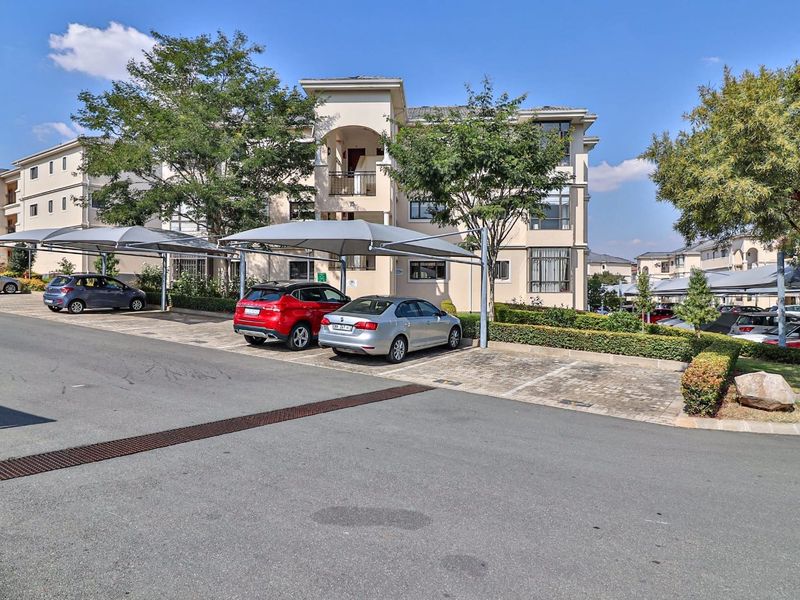 Exterior view of a multi-story apartment complex featuring paved parking areas with shade-cloth carports and landscaped garden borders.