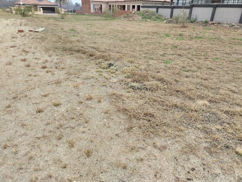 A view of a vacant residential plot featuring dry grass and sandy soil, with neighboring residential structures visible in the background.