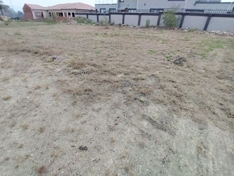 A vacant residential plot featuring dry grass and gravel terrain, enclosed by a perimeter wall with adjacent residential properties visible in the background.