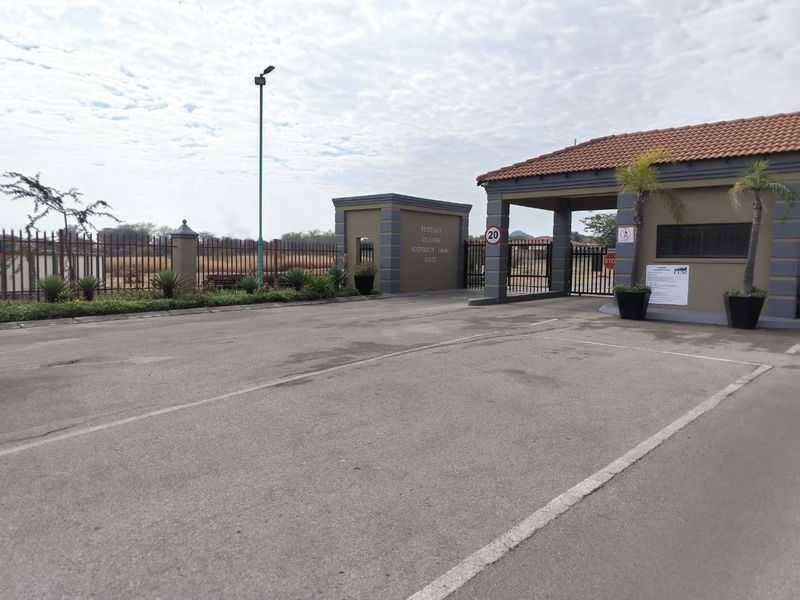 Entrance gate and security access point for Tuscany Village, featuring paved driveway, perimeter fencing, and tiled roof structure.