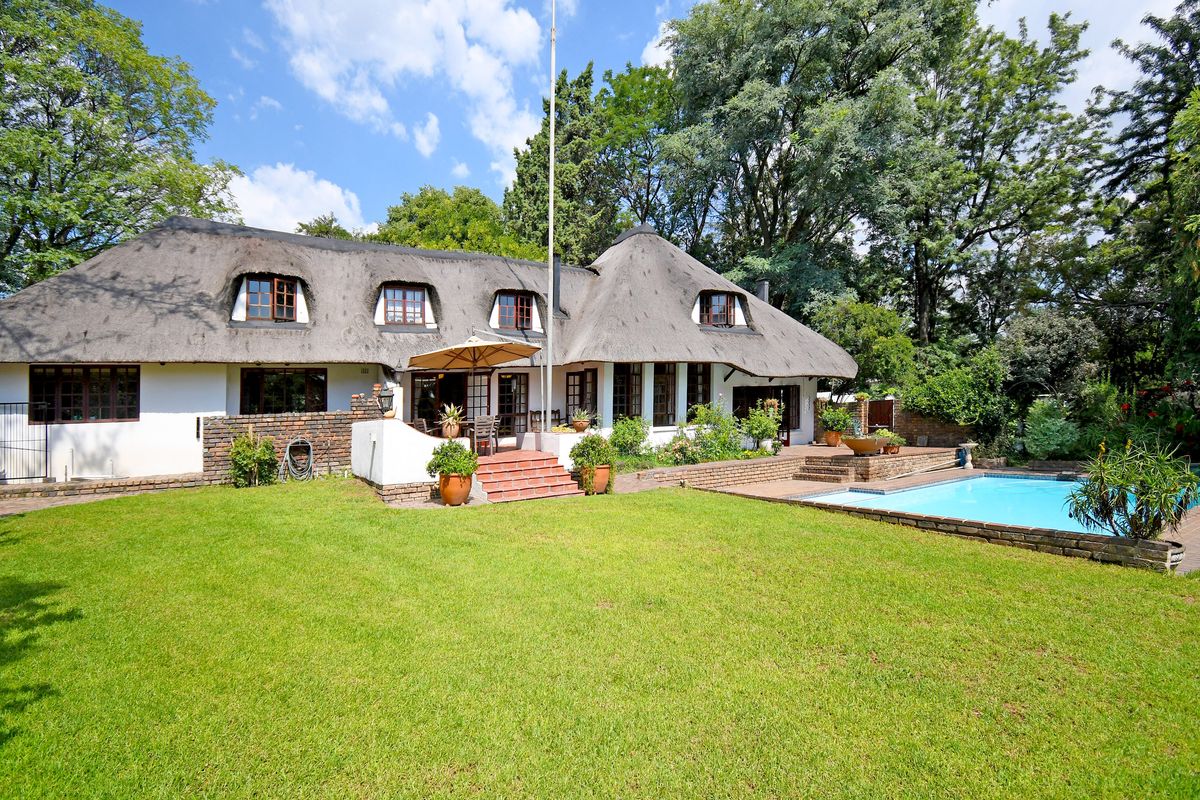 A rear view of a charming thatched-roof house featuring a spacious lawn, a swimming pool, and a paved patio area.