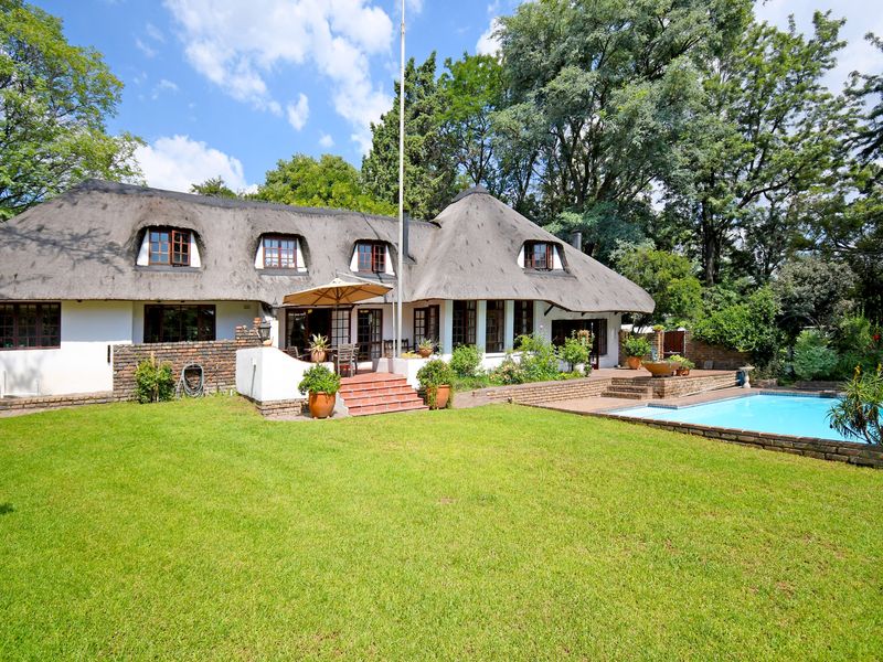 A rear view of a charming thatched-roof house featuring a spacious lawn, a swimming pool, and a paved patio area.