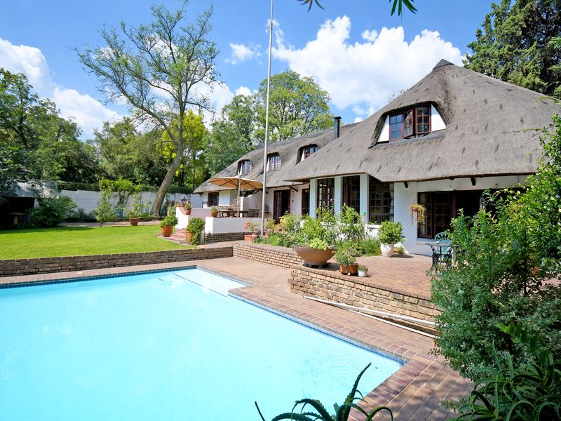 Rear view of a traditional thatched-roof house featuring a swimming pool, brick-paved patio area, and a well-maintained garden.