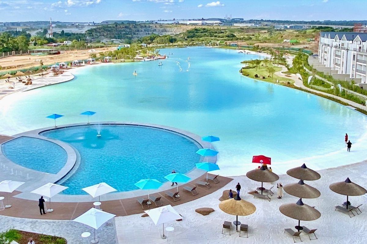 Aerial view of a large crystal lagoon and swimming pool area featuring sandy beaches, landscaped surroundings, and modern apartment buildings.