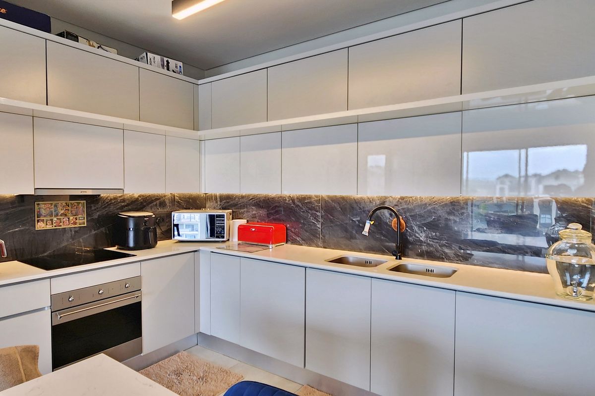 Modern kitchen featuring sleek white handleless cabinetry, integrated oven, induction hob, extractor fan, and a striking dark marble-effect splashback with under-cabinet lighting.