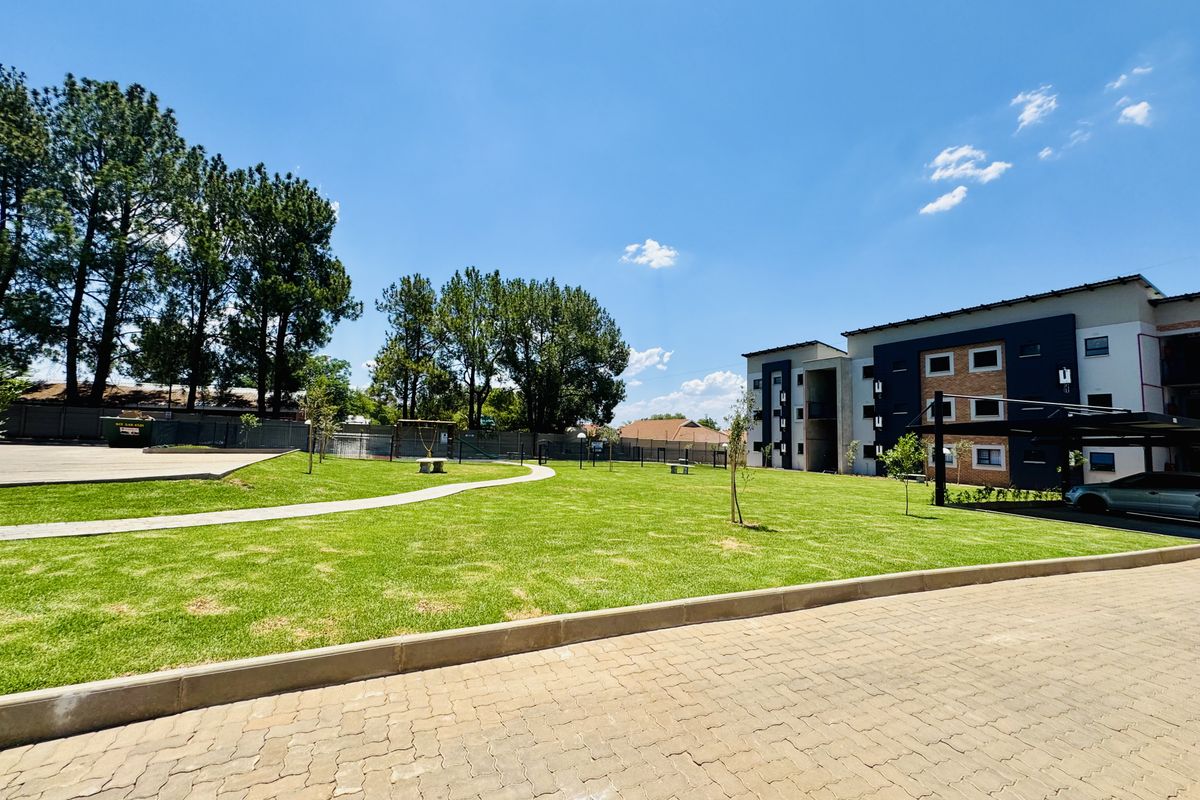 A well-maintained communal garden area featuring a paved walkway, manicured lawn, and modern apartment building exterior with covered parking bays.