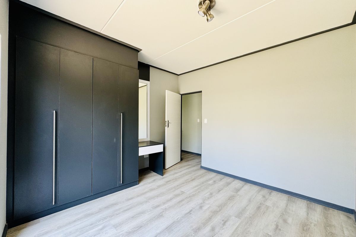 A bright, modern bedroom featuring built-in dark cabinetry with an integrated vanity and light wood-look laminate flooring.