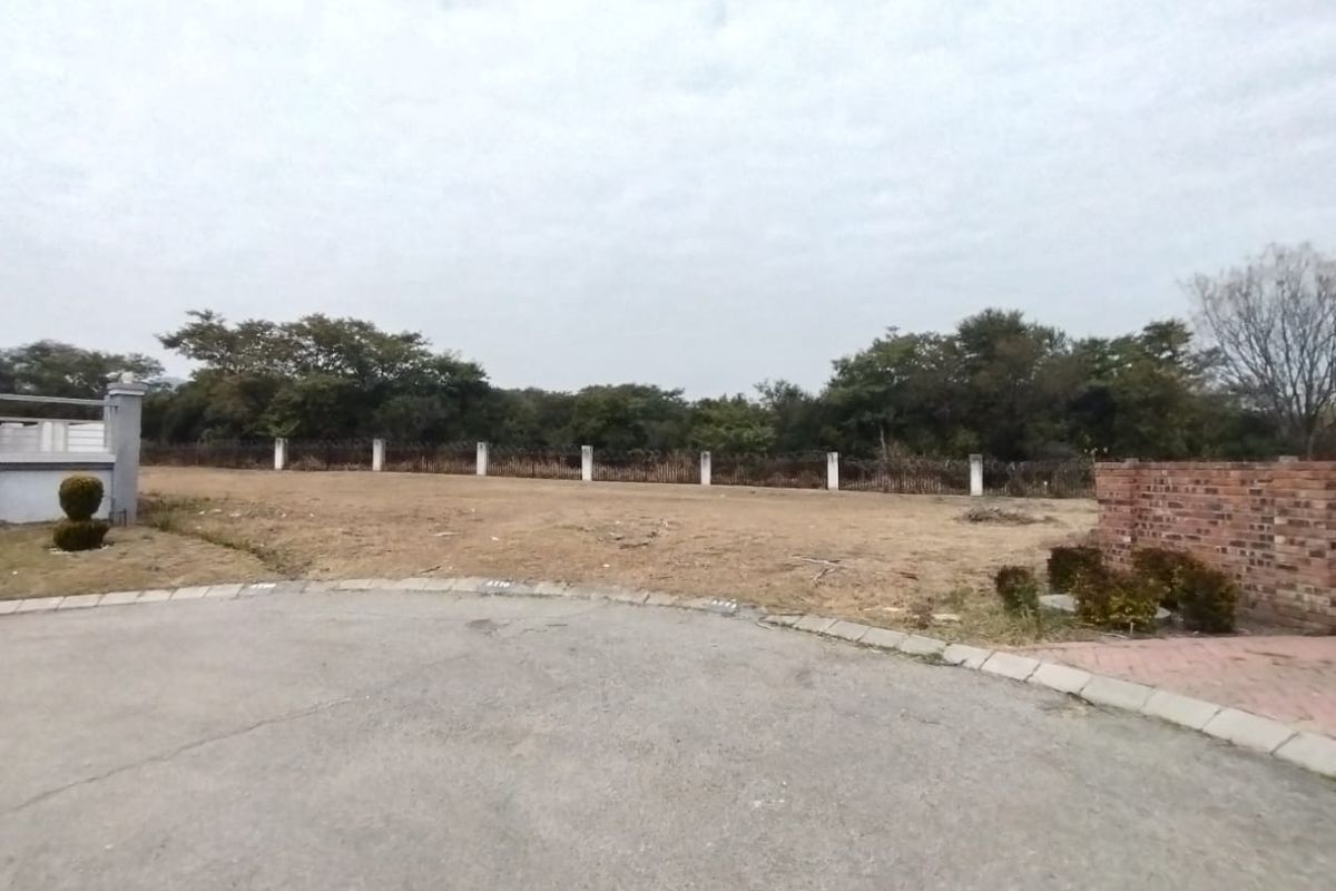 A vacant residential plot featuring a paved driveway entrance, perimeter brick walling, and established boundary fencing with mature trees in the background.