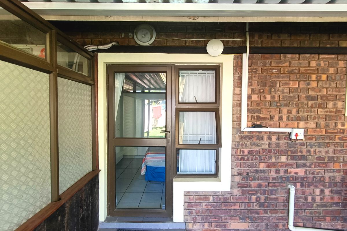 Exterior view of an apartment entrance featuring a brick facade, aluminium framed door, and window under a corrugated roof covering.