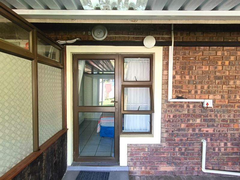 Exterior view of an apartment entrance featuring a brick facade, aluminium framed door, and window under a corrugated roof covering.