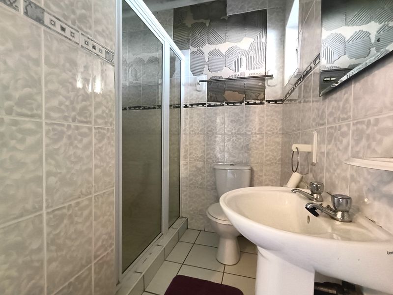 A bathroom featuring a pedestal basin, toilet, and shower enclosure, currently requiring repair due to missing wall tiles and exposed adhesive.