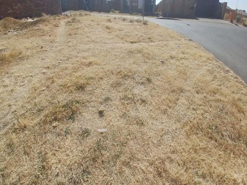A vacant residential plot of land featuring dry grass, situated adjacent to a paved street in a developed neighborhood.