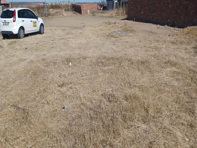 A vacant residential plot featuring dry grass, level terrain, and surrounding brick boundary walls in a developing neighborhood.