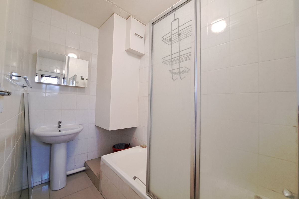 A functional bathroom featuring a pedestal basin, a built-in bathtub with a shower screen, and wall-mounted storage cabinetry with white tiled finishes.