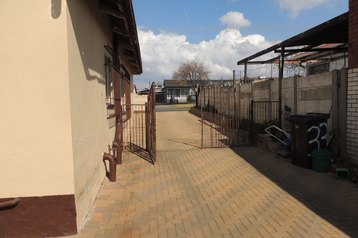 A paved side driveway area featuring a metal security gate, precast concrete boundary wall with razor wire, and an attached metal carport structure.