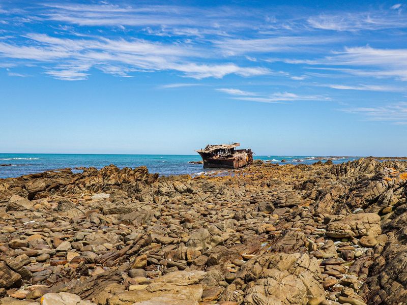 A scenic coastal view featuring a rocky shoreline and a shipwreck in the distance under a clear blue sky.
